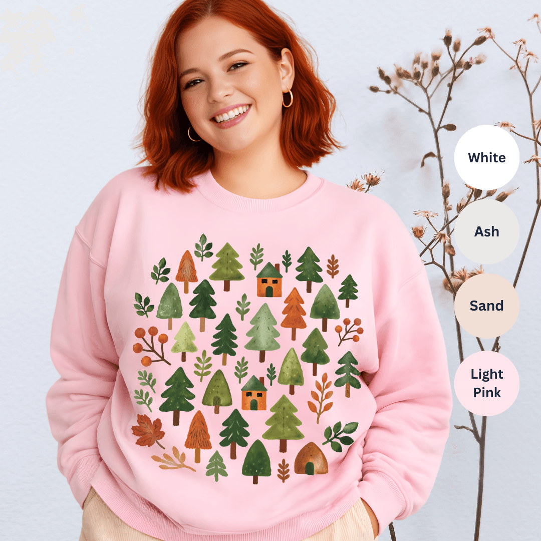 Light pink Gildan 18000 crewneck sweatshirt with cozy forest illustration of pine trees and cabins in autumnal greens and rusts