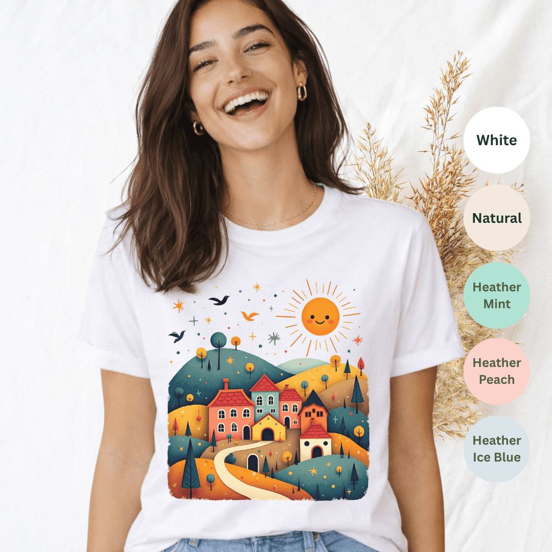 White Bella+Canvas 3001 t-shirt with sunny village folk art scene featuring colorful rooftops and rolling hills