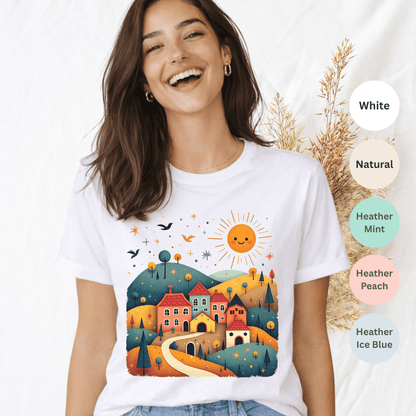 White Bella+Canvas 3001 t-shirt with sunny village folk art scene featuring colorful rooftops and rolling hills