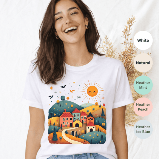 White Bella+Canvas 3001 t-shirt with sunny village folk art scene featuring colorful rooftops and rolling hills